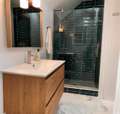 Modern bathroom with a wooden vanity, white countertop, rectangular mirror, and glass-enclosed shower with dark green tiles.